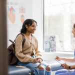 What's the difference between pediatrics and adolescent medicine? Young adult female patient smiles while listening to the nurse give an encouraging update regarding her medical exam